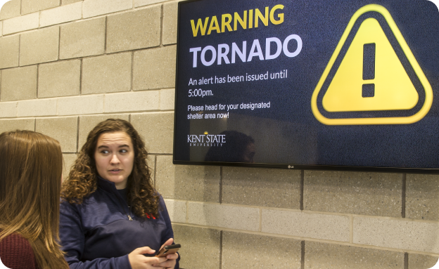 Two females standing next to a digital dispay about emergency alerts