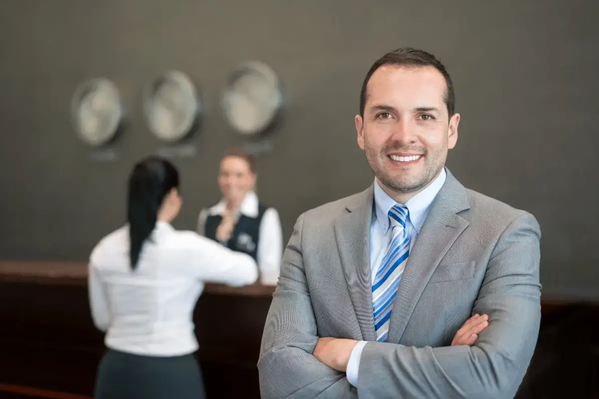 Man standing in a hotel with a concierge in background helping a guest
