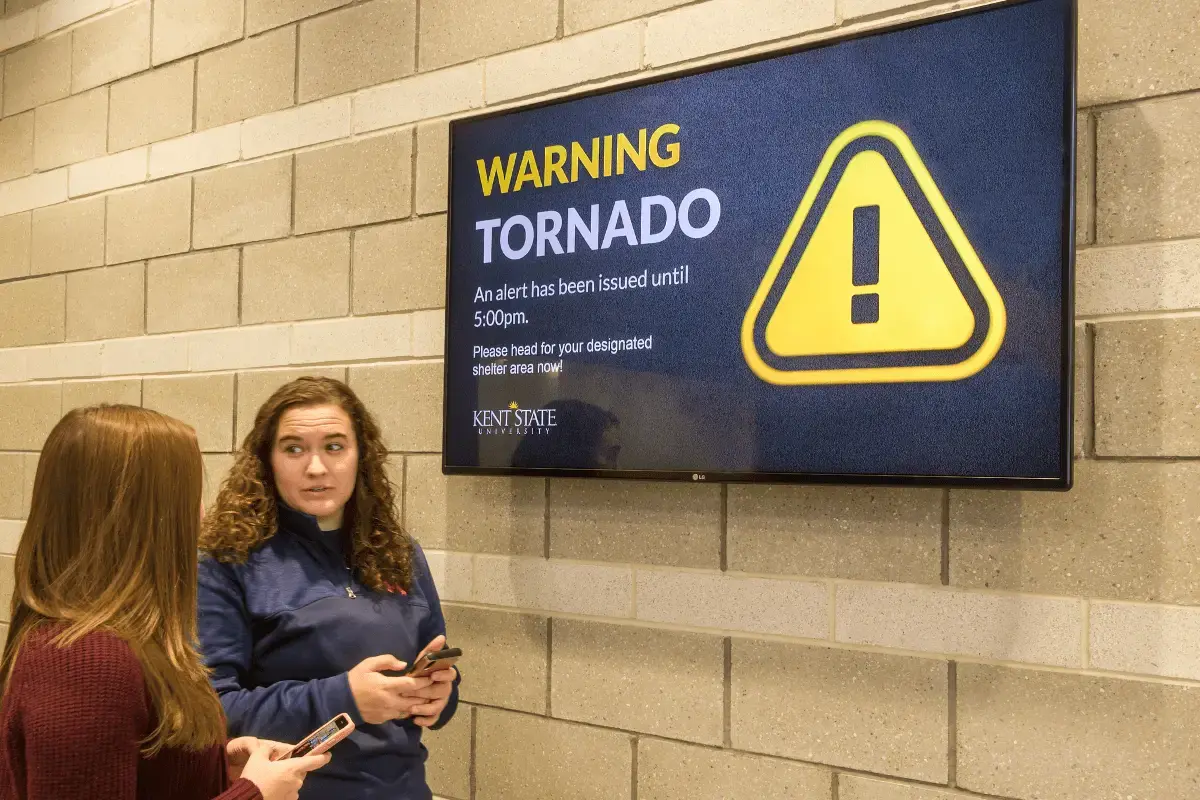 two female students standing next to a wall with emergency digital signage displayed on a TV